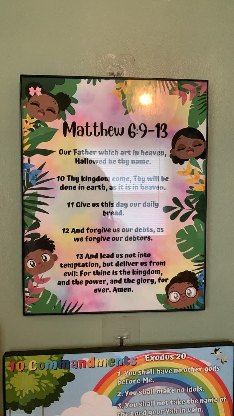 May include: A framed print featuring the text "Matthew 6:9-13" with a colorful floral and cartoon character design. The text includes a prayer. The frame is black.