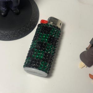 May include: A decorative lighter with a red button and a white base. The lighter is embellished with black and green crystals. The lighter is on a white surface.