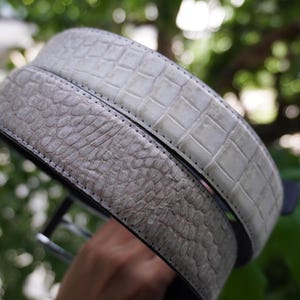 May include: Two textured leather belts in shades of gray. One belt has a crocodile pattern, and the other has a reptile-like texture. Both belts have black edges and stitching, suggesting a high-quality design.