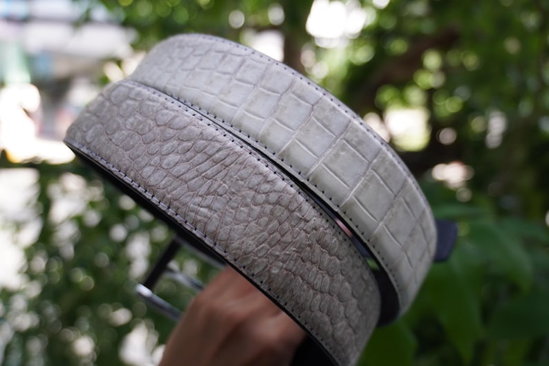 May include: Two textured leather belts in shades of gray. One belt has a crocodile pattern, and the other has a reptile-like texture. Both belts have black edges and stitching, suggesting a high-quality design.