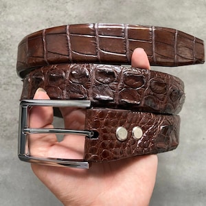 May include: A brown leather belt with a crocodile pattern and a silver buckle. The belt is folded in half and held in a hand.