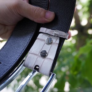 May include: Close-up of a black leather belt with a white alligator leather accent. The belt has a silver buckle and is held up by a hand. The belt is a fashion accessory.