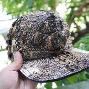 K&ouml;nnte beinhalten: Eine Baseballkappe mit einer strukturierten, reptilienartigen Optik. Die Kappe hat einen gebogenen Schirm und eine Krone mit einem schuppenartigen Muster in Braun- und Beiget&ouml;nen. Das Design der Kappe ahmt das Aussehen von Krokodil- oder Alligatorkaut nach.