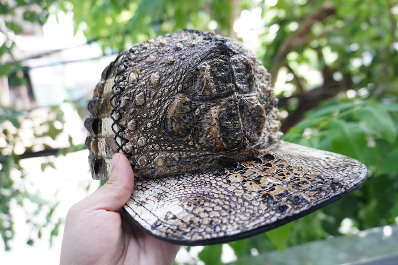 K&ouml;nnte beinhalten: Eine Baseballkappe mit einer strukturierten, reptilienartigen Optik. Die Kappe hat einen gebogenen Schirm und eine Krone mit einem schuppenartigen Muster in Braun- und Beiget&ouml;nen. Das Design der Kappe ahmt das Aussehen von Krokodil- oder Alligatorkaut nach.