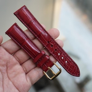 May include: A pair of red watch straps with a crocodile skin pattern. The straps have a gold-colored buckle and are held in a hand.
