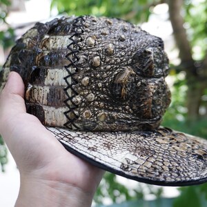 K&ouml;nnte beinhalten: Eine Baseballkappe mit einer strukturierten, reptilienartigen Optik. Die Kappe hat einen gebogenen Schirm und eine Krone mit einem Muster, das Krokodilhaut &auml;hnelt. Die Farbpalette umfasst Braun-, Beige- und Schwarzt&ouml;ne.