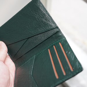 May include: Open, dark green leather wallet with multiple card slots. The wallet is held open, revealing the interior card slots and the textured leather. The wallet's interior is a lighter shade of green.
