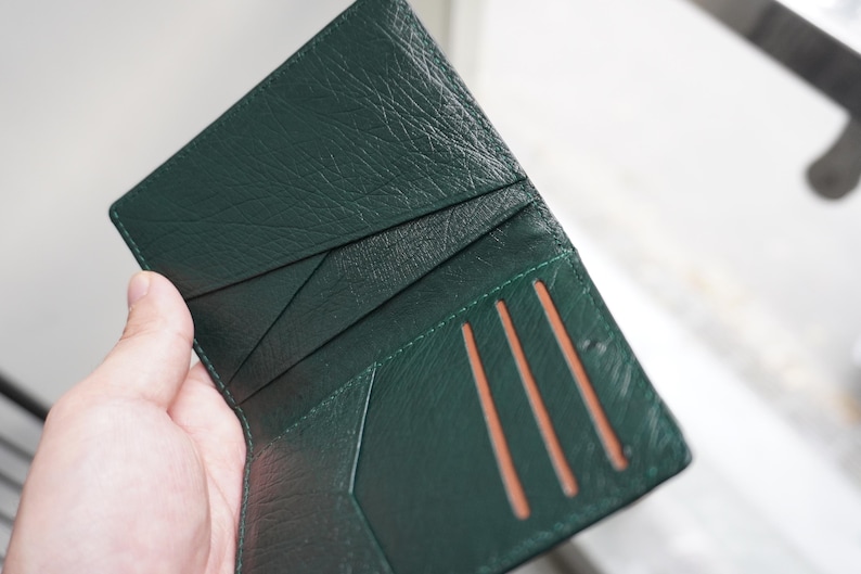 May include: Open, dark green leather wallet with multiple card slots. The wallet is held open, revealing the interior card slots and the textured leather. The wallet's interior is a lighter shade of green.