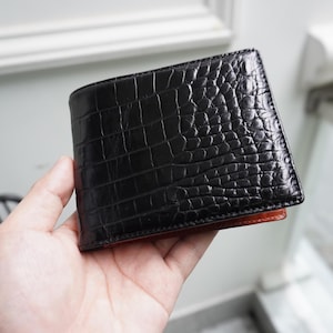 May include: A black leather wallet with a crocodile skin texture. The wallet is rectangular and has a bi-fold design. The interior is a reddish-brown color. The wallet is held in a person's hand.