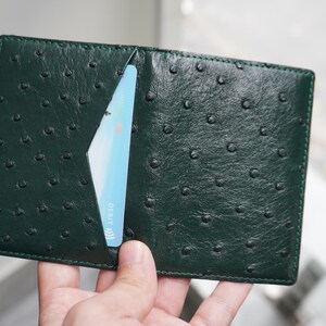 May include: Dark green leather wallet with a textured ostrich pattern. A light blue credit card is partially visible in a card slot. The wallet is open, showcasing its interior design.