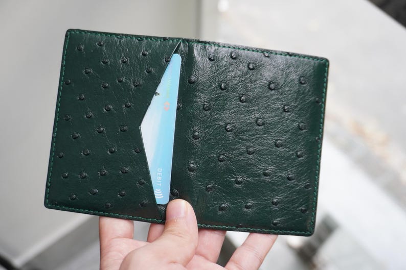 May include: Dark green leather wallet with a textured ostrich pattern. A light blue credit card is partially visible in a card slot. The wallet is open, showcasing its interior design.