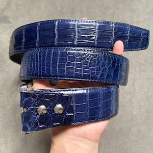 Peut inclure: Une ceinture en cuir d'alligator bleu avec une boucle argentée. La ceinture est pliée en trois et tenue dans une main.