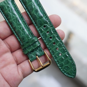 Puede incluir: Correa de reloj verde con un patrón de cocodrilo. La correa tiene una hebilla dorada y está hecha de un material texturizado. La correa del reloj se muestra sobre un fondo neutro.