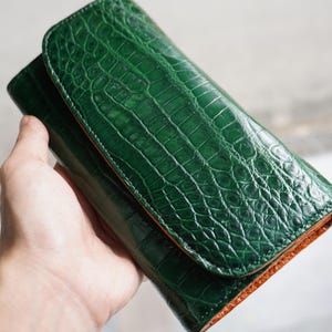 Op de afbeelding: Een groene leren portemonnee met een krokodillenleertextuur. De portemonnee is diep smaragdgroen met een contrasterend oranje interieur. De portemonnee wordt opengehouden en toont het interieur en de textuur van het leer.