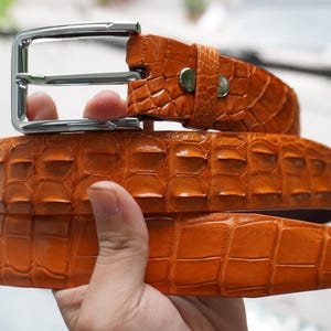 May include: An orange leather belt with a crocodile skin effect. The belt features a silver-coloured metal buckle. The belt is folded, displaying the detailed texture and colour. The belt is approximately 3.8 cm wide.