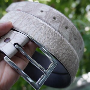 May include: A light gray belt with a textured, crocodile-like pattern. The belt has a silver-colored rectangular buckle and several holes for adjustment. The belt is made of a durable material and is suitable for everyday wear.