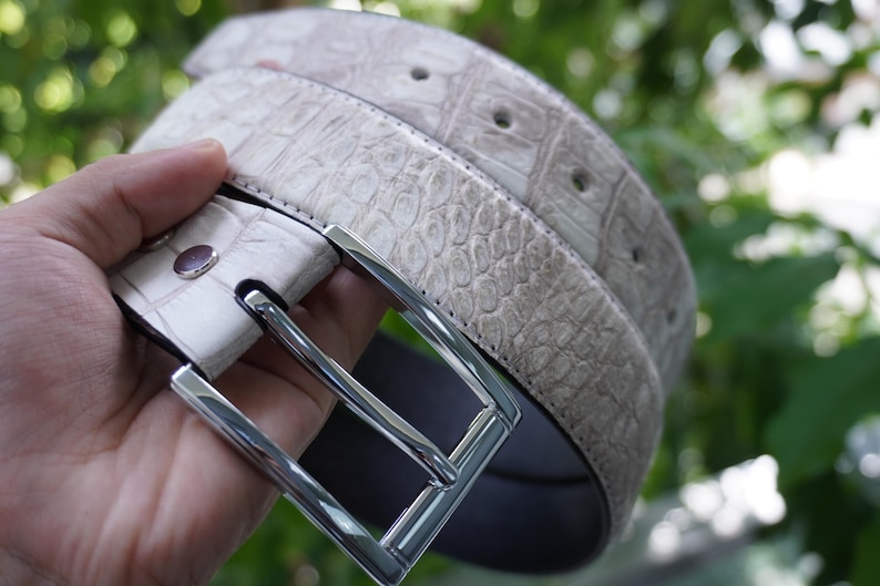 May include: A light gray belt with a textured, crocodile-like pattern. The belt has a silver-colored rectangular buckle and several holes for adjustment. The belt is made of a durable material and is suitable for everyday wear.