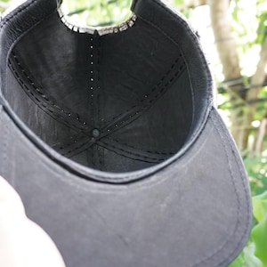 K&ouml;nnte beinhalten: Eine schwarze Baseballkappe mit gebogenem Schirm. Das Innere der Kappe ist sichtbar und zeigt ein dunkles Futter und N&auml;hte. Die Kappe wird hochgehalten, wobei eine Hand teilweise im Bild zu sehen ist. Der Hintergrund ist verschwommenes gr&uuml;nes Laub.