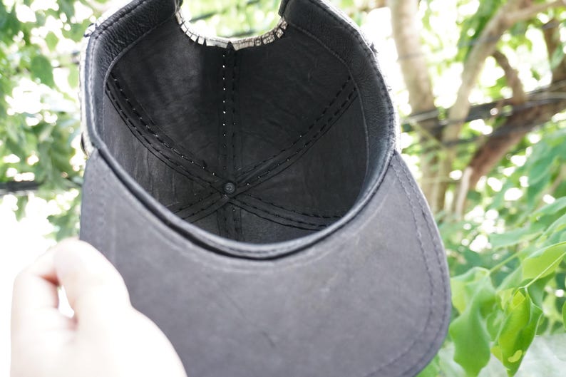 K&ouml;nnte beinhalten: Eine schwarze Baseballkappe mit gebogenem Schirm. Das Innere der Kappe ist sichtbar und zeigt ein dunkles Futter und N&auml;hte. Die Kappe wird hochgehalten, wobei eine Hand teilweise im Bild zu sehen ist. Der Hintergrund ist verschwommenes gr&uuml;nes Laub.