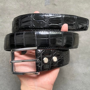 May include: Black alligator leather belt with a silver buckle. The belt is folded in half and held in a hand.