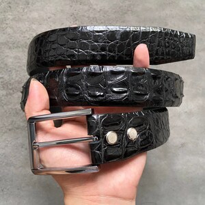 Pode incluir: Um cinto de couro preto com padrão de crocodilo e fivela prateada. O cinto está dobrado em três partes e segurado em uma mão.