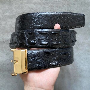 Peut inclure: Une ceinture en cuir d'alligator noir avec une boucle dorée. La ceinture est enroulée et tenue dans une main.