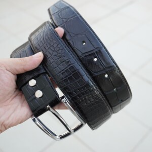 May include: Two black leather belts with a crocodile pattern. The belts are stacked on top of each other and have a silver buckle.
