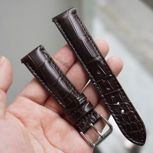 Peut inclure: Deux bracelets de montre marron foncé avec une texture peau de crocodile. L'un des bracelets a une boucle argentée. Les bracelets sont tenus dans la main d'une personne sur un fond flou.