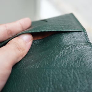 May include: Close-up of a dark green leather wallet. The wallet is open, revealing a glimpse of the interior. The leather has a textured appearance. The wallet is being held open by a hand.
