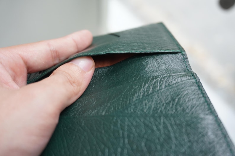 May include: Close-up of a dark green leather wallet. The wallet is open, revealing a glimpse of the interior. The leather has a textured appearance. The wallet is being held open by a hand.