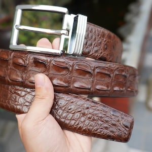 Könnte beinhalten: Ein brauner Ledergürtel mit geprägtem Krokodilmuster. Der Gürtel hat eine silberfarbene, rechteckige Schnalle. Der Gürtel wird in einer Hand gehalten, wodurch die Details des Leders und der Schnalle hervorgehoben werden.