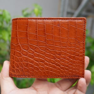 May include: A brown leather wallet with a crocodile skin texture. The wallet is held in a person's hand, showcasing its rectangular shape and detailed surface. The color is a warm, reddish-brown.