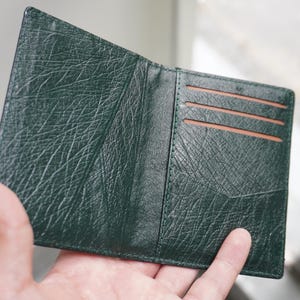 May include: Open, dark green leather wallet with a textured surface. The wallet has three card slots and a pocket. The interior is a lighter brown color. The wallet is held open by a hand.