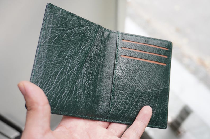 May include: Open, dark green leather wallet with a textured surface. The wallet has three card slots and a pocket. The interior is a lighter brown color. The wallet is held open by a hand.