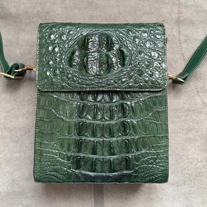 Op de afbeelding: Een donkergroene crossbody tas van krokodillenleer met een klep sluiting en een gouden gesp. De tas heeft een lange verstelbare band.