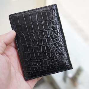 May include: A black, rectangular wallet with a textured, crocodile-like pattern. The wallet is held in a hand and appears to be made of leather. The wallet has a sleek, minimalist design.