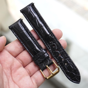 Puede incluir: Correas de reloj negras con un patrón texturizado similar al de un cocodrilo. Las correas tienen hebillas doradas y están diseñadas para sujetarse a la esfera del reloj. La piel parece ser de alta calidad.