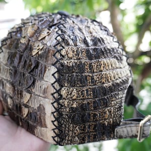 K&ouml;nnte beinhalten: Baseballkappe mit einem strukturierten, reptilienartigen Muster in Braun- und Schwarzt&ouml;nen. Die Kappe hat einen gebogenen Schirm und einen verstellbaren Riemen mit einer goldfarbenen Schnalle. Die N&auml;hte sind schwarz.