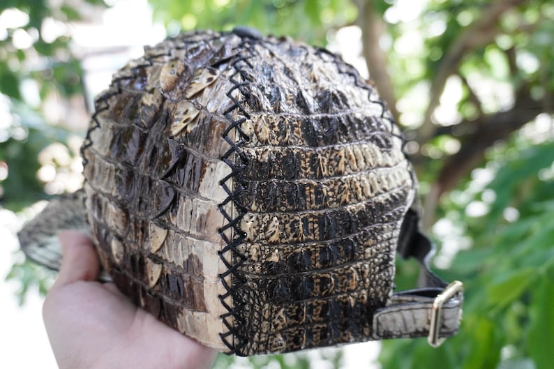K&ouml;nnte beinhalten: Baseballkappe mit einem strukturierten, reptilienartigen Muster in Braun- und Schwarzt&ouml;nen. Die Kappe hat einen gebogenen Schirm und einen verstellbaren Riemen mit einer goldfarbenen Schnalle. Die N&auml;hte sind schwarz.