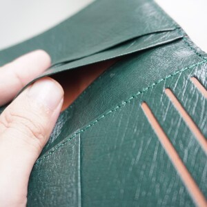 May include: Close-up of a dark green leather wallet with visible stitching and card slots. The wallet is open, revealing a brown interior. The leather has a textured appearance.