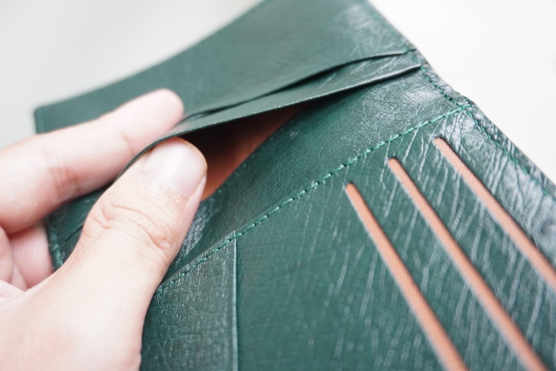May include: Close-up of a dark green leather wallet with visible stitching and card slots. The wallet is open, revealing a brown interior. The leather has a textured appearance.