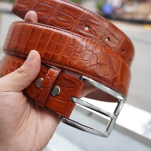 May include: A brown leather belt with a crocodile skin texture. The belt has a silver-colored buckle and two silver rivets. The belt is held in a hand, showcasing its color and texture. The belt is a fashion accessory.