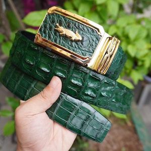 Op de afbeelding: Een groene leren riem met een gouden gesp. De gesp heeft een gouden alligator-embleem en een rechthoekig frame. De riem heeft een textuur met krokodillenpatroon. De riem wordt in een hand gehouden.