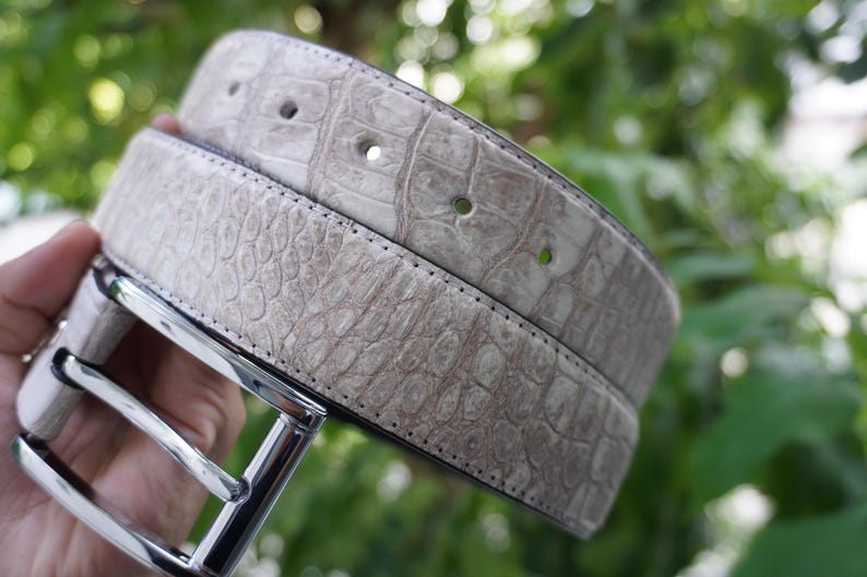 May include: A light gray belt with a textured, crocodile-like pattern. The belt has a silver-colored buckle and several holes for adjustment. The background is blurred green foliage.