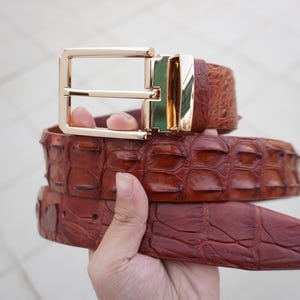 Puede incluir: Un cinturón de cuero marrón con estampado de cocodrilo y hebilla dorada.