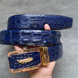 Pode incluir: Um cinto de couro de jacaré azul com uma fivela dourada com design de jacaré.