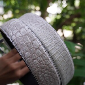 May include: A light gray belt with a textured, crocodile-like pattern. The belt is made of a durable material and features a silver-colored buckle. The design is suitable for both casual and formal wear.