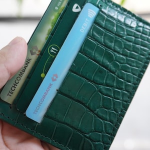 May include: A dark green wallet with a crocodile skin effect. Several credit cards are visible, including a light blue debit card. The wallet is held in a hand.