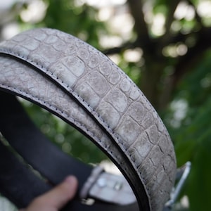 May include: A close-up of a gray belt with a textured, crocodile-like pattern. The belt has a silver buckle and a dark gray interior. The belt is made of a durable material and is suitable for everyday wear.