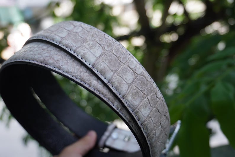 May include: A close-up of a gray belt with a textured, crocodile-like pattern. The belt has a silver buckle and a dark gray interior. The belt is made of a durable material and is suitable for everyday wear.